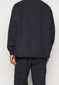 Black lightweight jacket with a relaxed fit, elastic cuffs, and a high collar. The back features minimal detailing and a smooth texture.