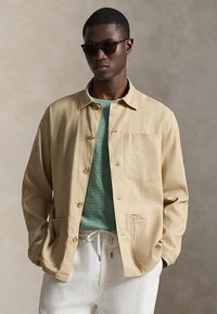 Άνδρας που φοράει σκούρα γυαλιά ηλίου, μπεζ σακάκι με κουμπιά πάνω από πράσινο πουλόβερ και λευκό παντελόνι με κορδόνι, στέκεται με τα χέρια στις τσέπες.