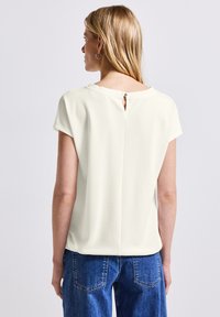 Haut blanc texturé avec une ouverture en forme de trou de serrure à l'arrière, avec des manches courtes et une coupe décontractée, associé à un pantalon en denim bleu.