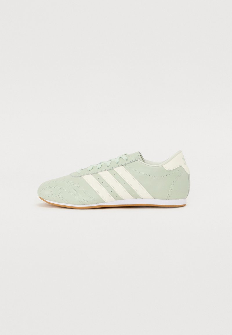 Zapatilla deportiva verde claro con rayas blancas, punta redonda, textura perforada y suela de goma, con un diseño minimalista y cierre de cordones.