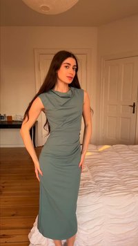 Γυναίκα με μακριά σκούρα μαλλιά που φοράει γκρι εφαρμοστό μίντι φόρεμα χωρίς μανίκια, στέκεται σε ένα υπνοδωμάτιο με ξύλινο πάτωμα και λευκά κλινοσκεπάσματα.