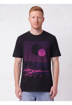 Homme aux cheveux clairs portant un T-shirt noir avec un paysage urbain violet et rose et deux grandes formes circulaires au-dessus.