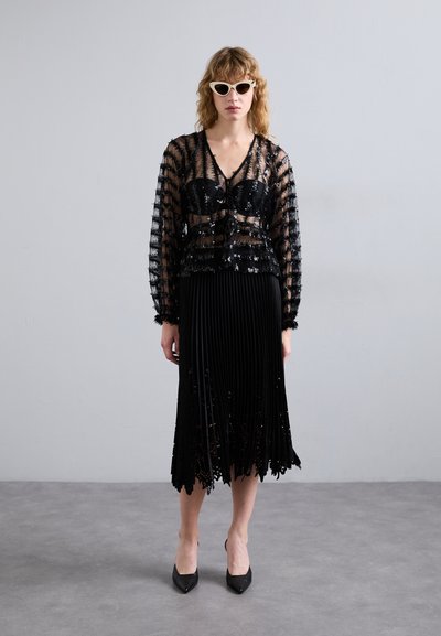 Läpinäkyvä musta pitkähiha toppi, jossa on paljettidetaljering, kombinert med plissert svart skjørt. Har spisse svarte hæler og hvite katteøye-solbriller.