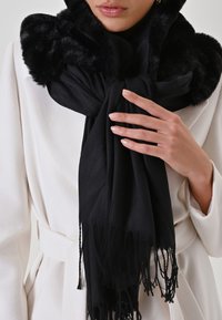 Sciarpa nera con morbido bordo in pelliccia sintetica, texture tessuta e estremità frangiate, appoggiata attorno al collo. Indossata con un cappotto bianco.