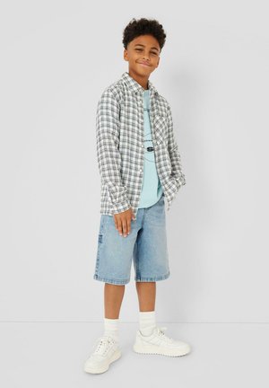 Garçon debout et souriant, portant une chemise à carreaux blanche par-dessus un t-shirt graphique bleu clair, un short en denim, des chaussettes blanches et des baskets blanches.