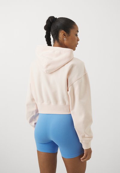 Haut à capuche rose pastel coupé, avec un cordon de serrage et un ourlet côtelé, associé à un short bleu uni, mettant en avant une coupe décontractée et une texture douce.