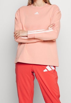 Vrouw met een lichtroze Adidas-trui met witte strepen op de mouwen, armen over elkaar, en felrode Adidas-broek met logo op de dij.