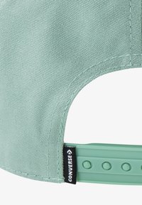 Gaiši zaļa auduma cepure ar fakturētu virsmu, regulējamu plastmasas siksniņu un melnu "Converse" uzrakstu sānos.