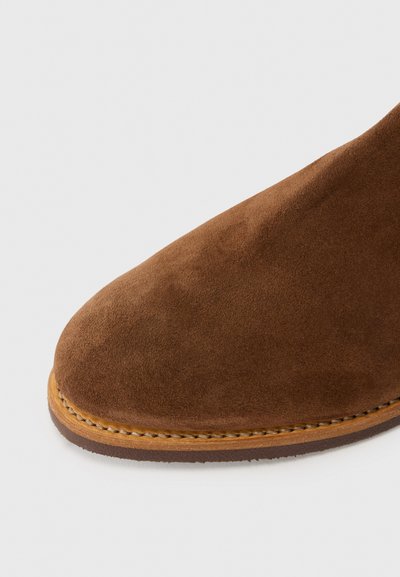 Botină din suede maro, cu vârful rotund, având o talpă din piele cosută și o textură fină, pe un fundal alb.