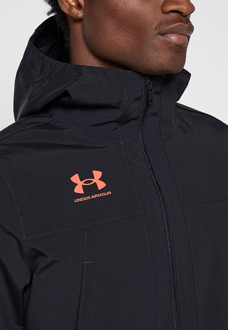 Veste imperméable noire avec un col haut, fermeture éclair, et logo Under Armour orange sur la poitrine gauche. Texture lisse et design extérieur.