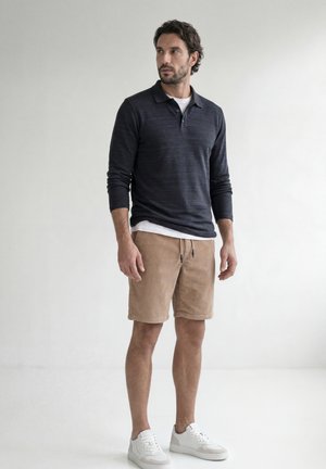 Mand med mørkt hår og skæg iført en navy langærmet polo, hvid undershirt, beige shorts og hvide sneakers, stående mod en ensfarvet lys baggrund.
