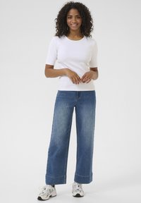 Camiseta blanca de algodón con mangas cortas, combinada con unos jeans de mezclilla azul de tiro alto y pierna ancha. El calzado incluye zapatillas blancas.