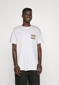 Usmívající se mladý muž v bílém tričku s barevným abstraktním nášivkem na hrudi a černými kalhotami, stojící na jednobarevném pozadí.