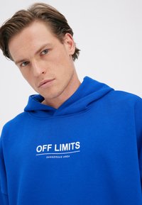 Mladý muž se světlou pletí a hnědými vlasy, který má na sobě modrou mikinu s bílým nápisem "OFF LIMITS" a "DANGEROUS AREA" na bílém pozadí.
