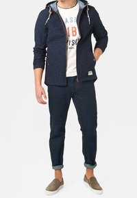 Marinblå zip-jacka med huva och ljusblå foder, kombinerad med en vit t-shirt med tryck, mörka jeans och gråa slip-on skor.