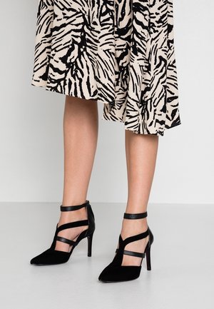 Des jambes portant une jupe zébrée noire et beige associée à des escarpins noirs à talons hauts avec des brides au niveau des chevilles et des découpes.