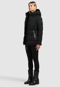 Žena stojící v černé péřové bundě s kapucí, černých legínách a černých kožených botách do poloviny lýtek na jednobarevném pozadí.