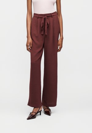 Γυναίκα που φοράει καφέ παντελόνι με ευρύ μπατζάκι και ψηλή μέση με ζώνη που δένει από το ίδιο ύφασμα, σε συνδυασμό με καφέ μυτερά παπούτσια με μικρό τακούνι, στέκεται μπροστά σε απλό φόντο.