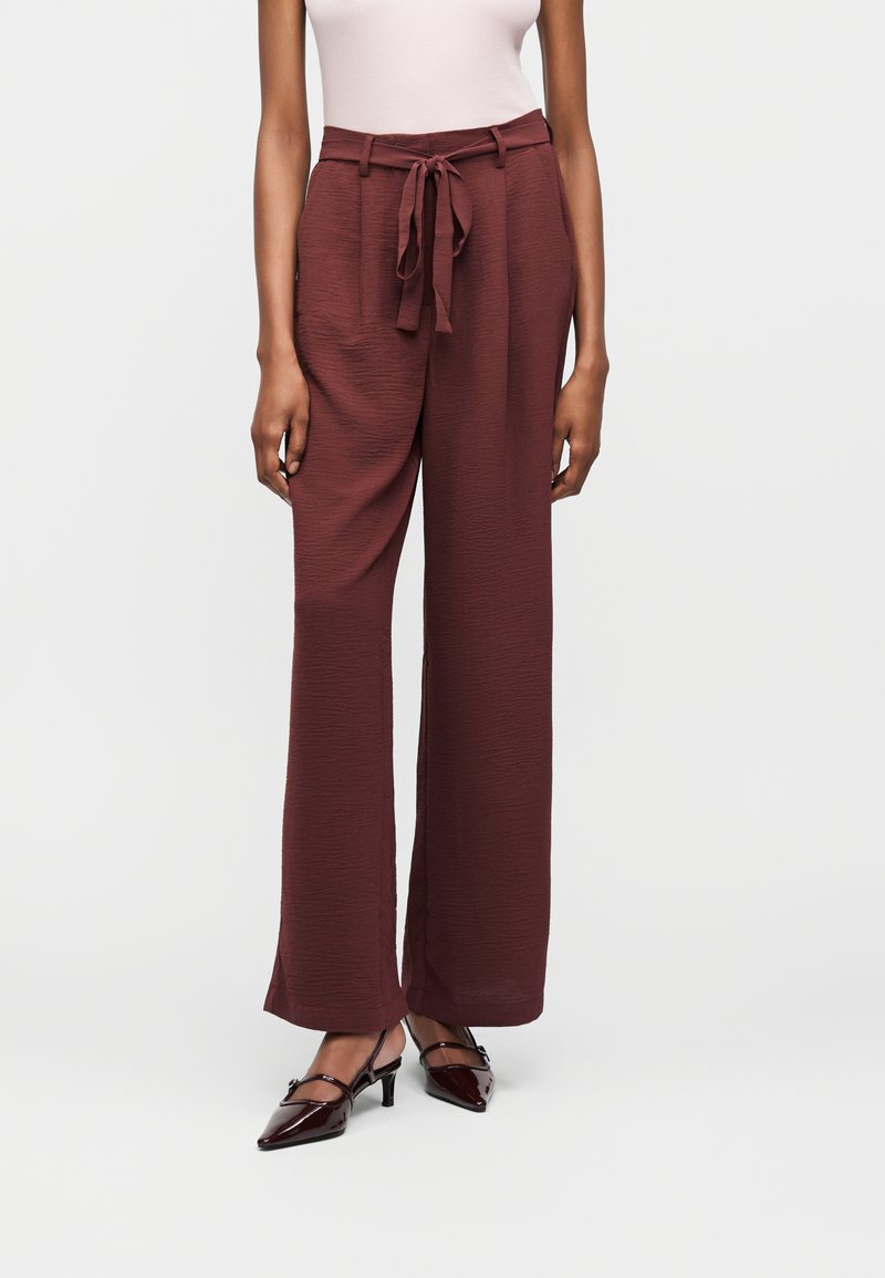 Γυναίκα που φοράει καφέ παντελόνι με ευρύ μπατζάκι και ψηλή μέση με ζώνη που δένει από το ίδιο ύφασμα, σε συνδυασμό με καφέ μυτερά παπούτσια με μικρό τακούνι, στέκεται μπροστά σε απλό φόντο.