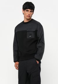 Sweatshirt negru cu texturi variate, având o secțiune superioară netedă și un buzunar tip patch. Pantaloni negri vizibili, menținând un aspect monocrom.