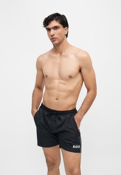 Homme torse nu aux cheveux foncés portant un short noir avec le logo BOSS, debout les mains dans les poches devant un fond blanc uni.