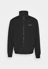 Czarna kurtka zapinana na zamek, wykonana z gładkiego, lekkiego materiału. Posiada wysoki kołnierz, elastyczne mankiety, boczne kieszenie oraz małe logo na piersi.