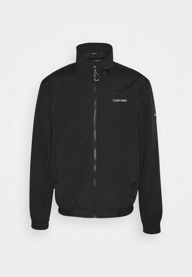 Czarna kurtka zapinana na zamek, wykonana z gładkiego, lekkiego materiału. Posiada wysoki kołnierz, elastyczne mankiety, boczne kieszenie oraz małe logo na piersi.