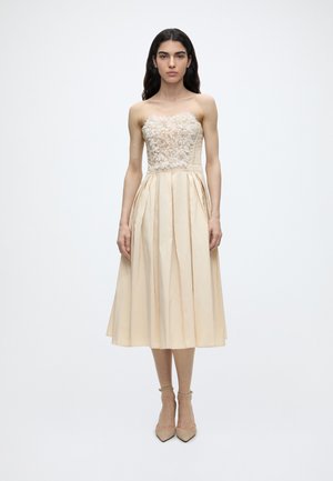 Femme portant une robe midi beige sans bretelles avec des détails floraux sur le corsage et une jupe plissée, debout devant un fond clair uni.