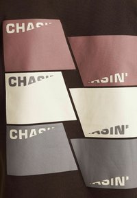 Grafische t-shirt met rechthoekige patches in gedempte kleuren (roze, wit, grijs) met de tekst "CHASIN'"; tegen een donkere achtergrond.