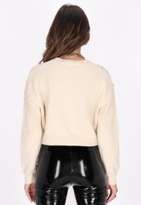 Cremiger Pullover mit strukturierten, offen gestrickten Akzenten an den Ärmeln und einem cropped Saum; kombiniert mit glänzenden, schwarzen, hochtaillierten Hosen.