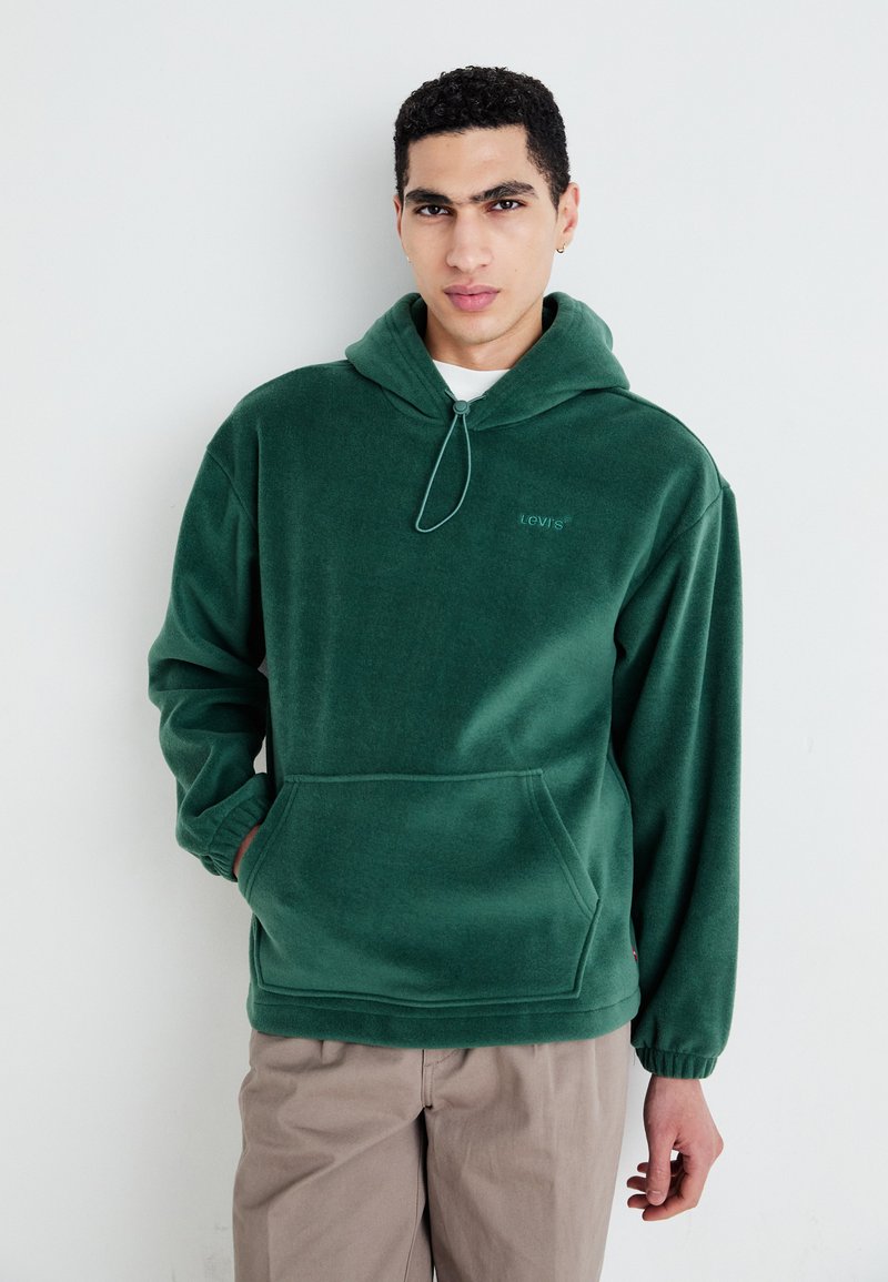 Grønn fleece hettegenser med snorhette, frontlomme og subtilt brodert logo. Myk tekstur og avslappet passform.