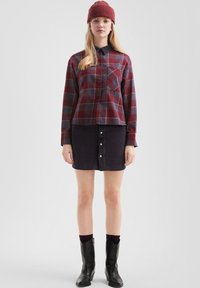 Geruite button-up blouse in bordeaux en donkergrijs, gecombineerd met een donkere corduroy rok met witte knopen, zwarte cowboylaarzen en een gebreide beanie.