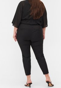 Svarta, figurnära byxor med en slät textur och avsmalnade ben, tillsammans med en lätt, genomskinlig svart topp med vertikala ränder.