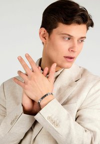 Bracciale d'argento con un intricato motivo a spina di pesce, indossato al polso di una persona in un compleanno beige chiaro con una finitura testurizzata e dettagli con bottoni.