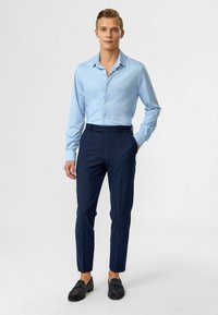 Lichtblauwe overhemng met lange mouwen, gecombineerd met donkerblauwe slim-fit broek en zwarte suède instappers. Eenvoudig, schoon ontwerp.