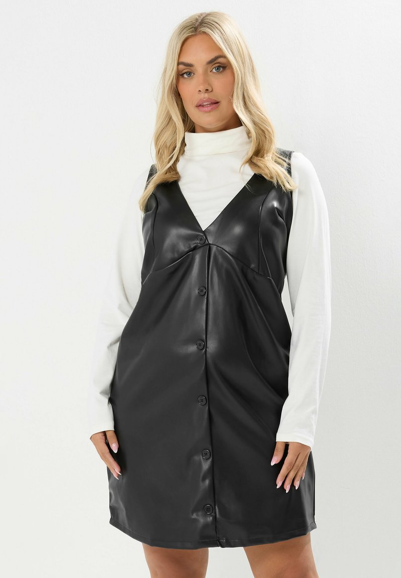 Abito in ecopelle nera con scollatura a V, design senza maniche, chiusura frontale con bottoni e vestibilità ampia, indossato sopra un dolcevita bianco.