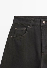 Zwarte spijkerbroek van denim met een hoge taille, voorzien van koperen klinknagels en contrasterende stiksels op zakken en naden. Platte tailleband met knoopsluiting.