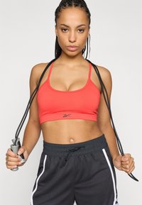 Κόκκινο αθλητικό σουτιέν με λεπτές τιράντες, μαύρη επωνυμία, σε συνδυασμό με μαύρη βερμούδα με λευκές ρίγες. Κράτημα σχοινιού άλματος με το ένα χέρι.