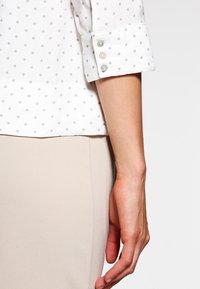 Vit skjorta med grå prickar, med uppvikt ärm som har tre knappar, i kombination med en beige botten. Slät textur.