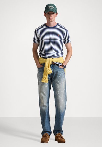 Noor mees kannab rohelist "Hudson Valley" mütsi, sinist ja valget triibulist särki, kollast kampsunit, mis on tal vööl, kulunud teksaseid ja pruune kingi.