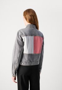 Persoon met lang bruin haar, gekleed in een grijze spijkerjacket met een grote witte en rode rechthoekige patch op de rug, en zwarte broek.