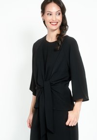 Top nero con vita annodata, maniche a tre quarti e una texture liscia. Design semplice con scollatura rotonda e tessuto stratificato.