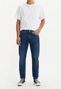 Personne portant un t-shirt blanc, un jean bleu, des baskets noires et blanches, et un collier chaîne argenté, debout devant un fond blanc uni.