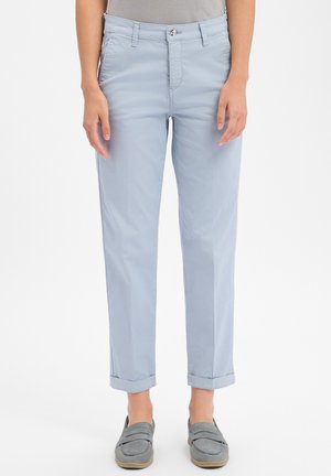 Personne portant un pantalon bleu clair retroussé et des chaussures grises sans lacets, debout devant un fond blanc uni.