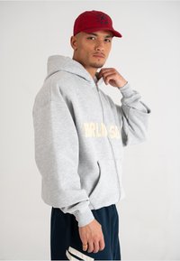 Felpa grigia con zip e logo frontale, dotata di due tasche laterali. Indossata con pantaloni blu navy con strisce bianche. Un cappello rosso completa il look.