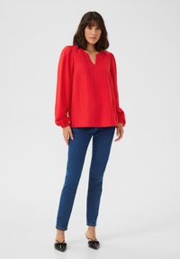 Haut rouge à manches longues avec un décolleté en V et un tissu texturé, associé à un jean slim bleu et des ballerines noires à bout pointu.