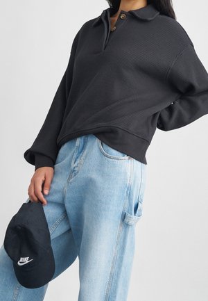 Persona vestida con un polo negro de manga larga con textura y jeans azul claro de talle alto, sosteniendo una gorra negra de Nike a un lado.