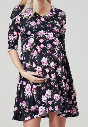 Raskaana oleva nainen mustassa kukkamekossa, jossa on vaaleanpunaisia ja valkoisia kukkia, pitäen vatsastaan hellästi yhdellä kädellä.