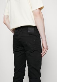 Schwarze Slim-Fit-Jeans mit einem Lederlabel am Bund und Gesäßtaschen mit genähten Details. Der Stoff wirkt glatt.