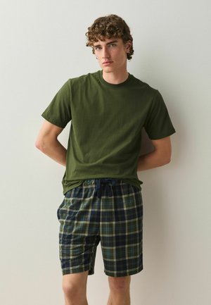 Junger Mann mit lockigem Haar, der ein dunkelgrünes T-Shirt und marineblaue karierte Shorts trägt, steht vor einem schlichten hellen Hintergrund.
